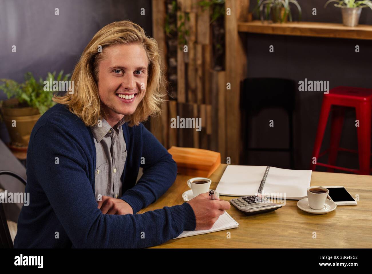 Uomo che scrive in un notebook con penna usando la calcolatrice al tavolo del caffè con tazze da caffè e smartphone Foto Stock
