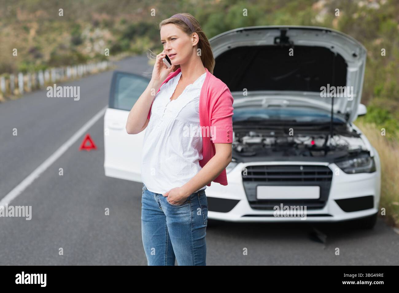 Donna della metà degli anni '30 che chiama sullo smartphone accanto alla berlina con il triangolo di avvertimento sollevato sul cofano sull'autostrada rurale Foto Stock