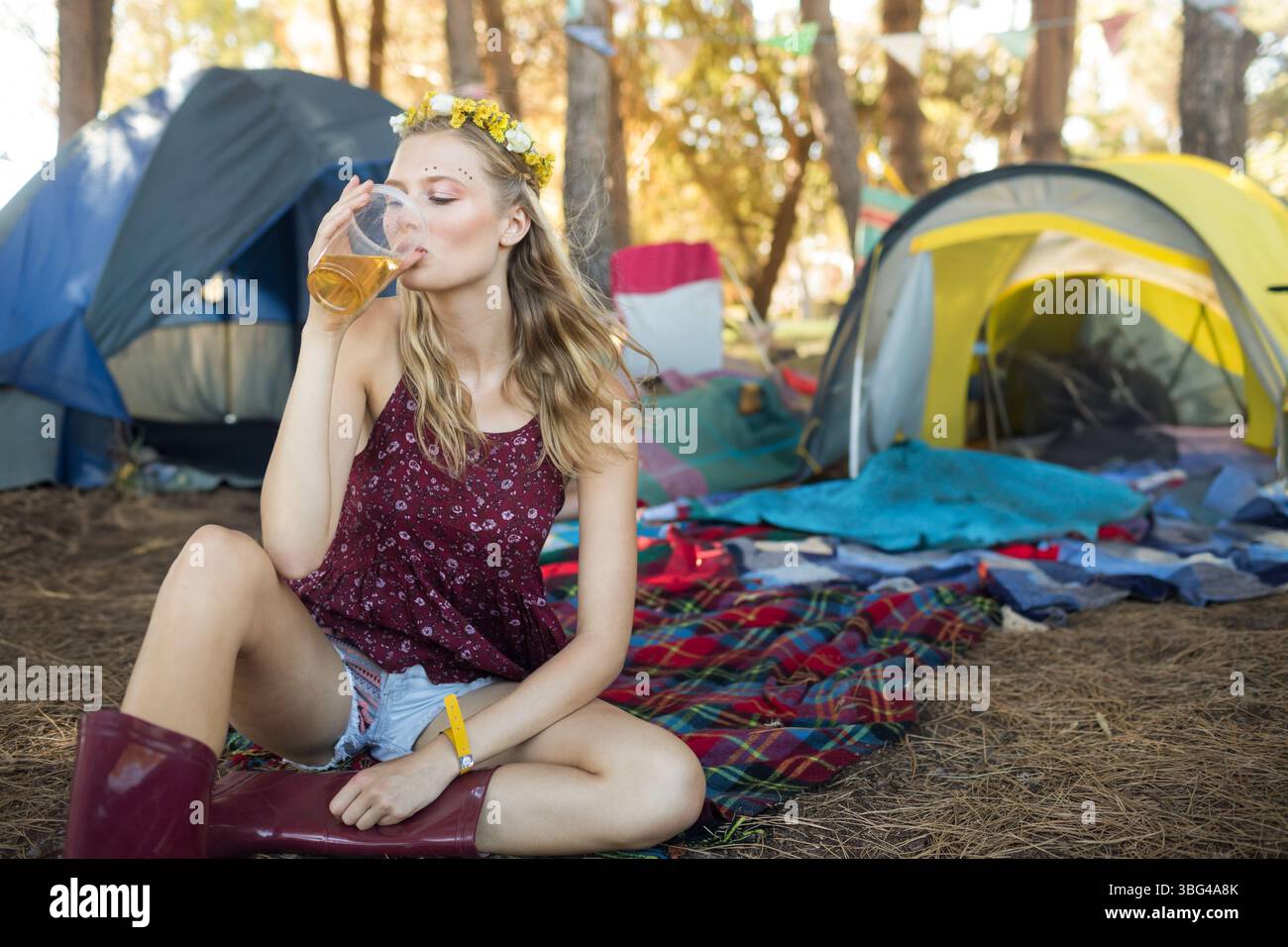 Donna seduta su una coperta a quadri al campo forestale con una corona floreale gialla che regge una tazza di plastica Foto Stock