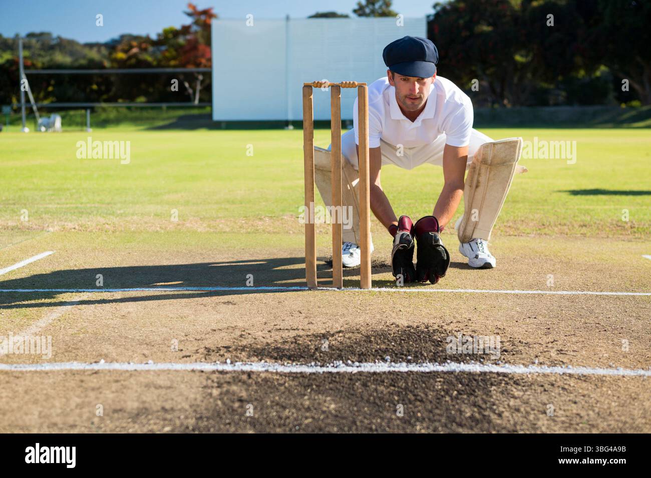 Guardino di wicket maschio che indossa un kit bianco accovacciato dietro i ceppi sui guanti da campo con schermo visivo Foto Stock