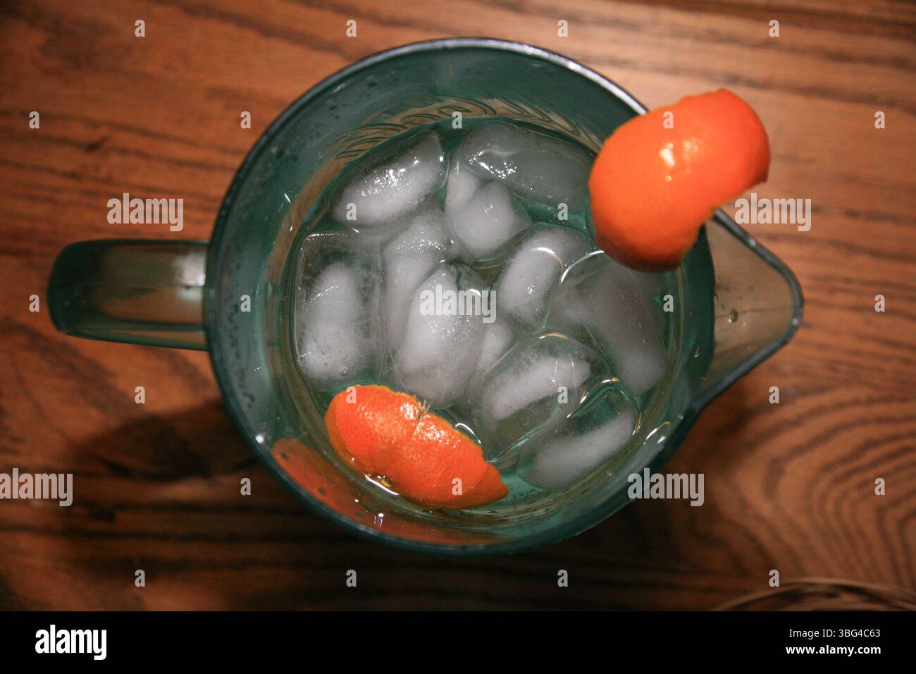 Vista dall'alto verso il basso di una caraffa di vetro piena di acqua ghiacciata e decorazioni di buccia d'arancia, adagiata su un tavolo di legno, un concetto di bevanda rinfrescante e vivace Foto Stock