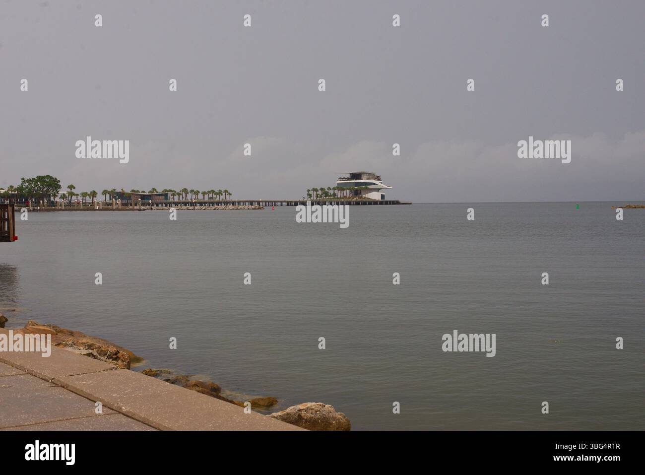 Molo di legno che si affaccia su un tranquillo paesaggio costiero in una giornata nuvolosa. A St. Petersburg, Florida. Il tranquillo molo in legno si estende con rocce in primo piano Foto Stock