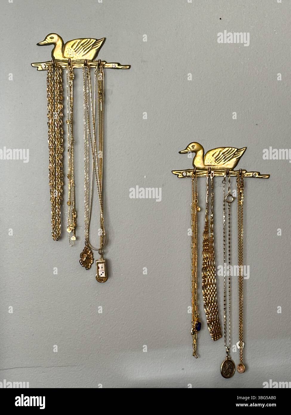Gli appendiabiti decorativi in ottone a forma di anatra espongono ordinatamente una varietà di collane e catene d'oro su una parete dipinta di grigio. Foto Stock