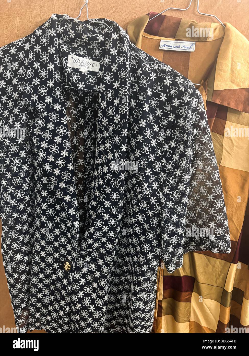 Una blusa YouWon nera con motivo floreale ricamata con spacco e una camicia a scacchi Anna e Frank marrone e oro su uno sfondo in legno. Foto Stock