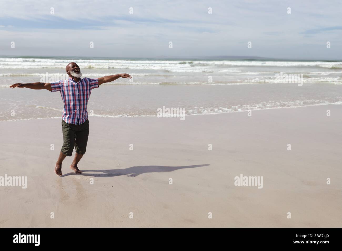 Uomo afro-americano che si allunga le braccia sulla spiaggia indossando pantaloni arrotolati a piedi nudi con le onde che rotolano Foto Stock