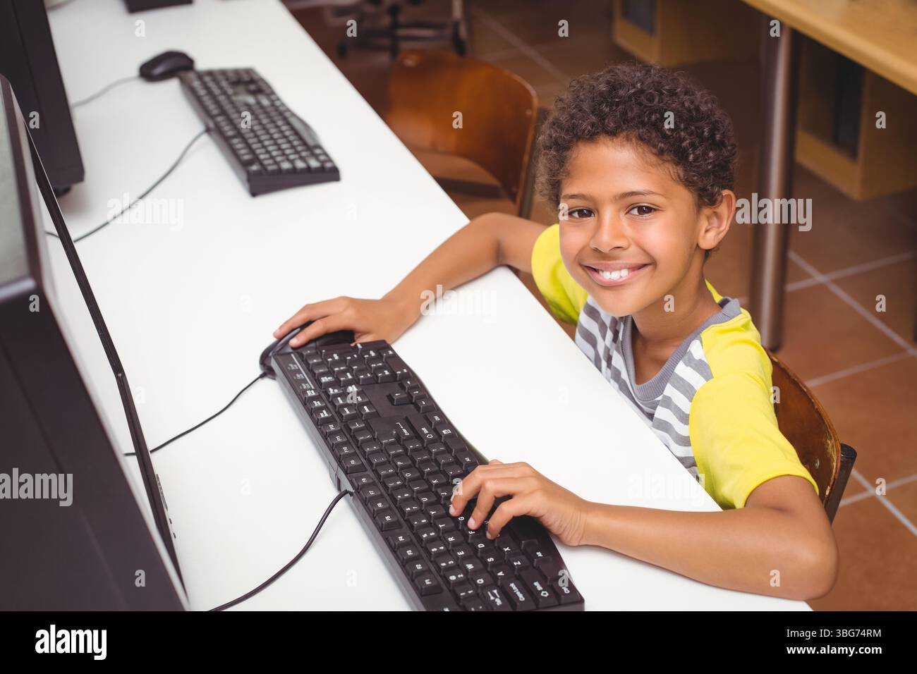 Ragazzo afroamericano sorridente e seduto alla scrivania del laboratorio di computer della scuola usando monitor e spazio di copia Foto Stock