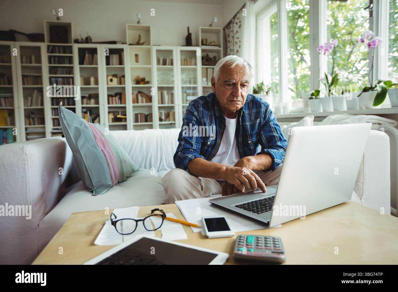Uomo che lavora su un computer portatile al tavolino da caffè in legno nel soggiorno, circondato da libri e orchidee Foto Stock