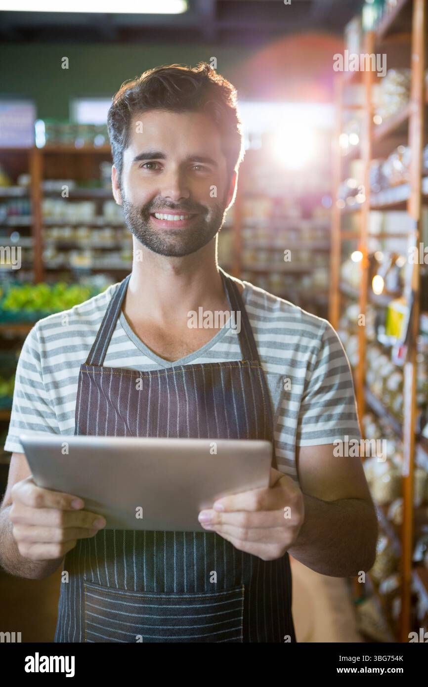 Impiegato maschio adulto che indossa un grembiule, tiene una compressa, controlla il magazzino nella corsia della spesa con ripiani in legno Foto Stock