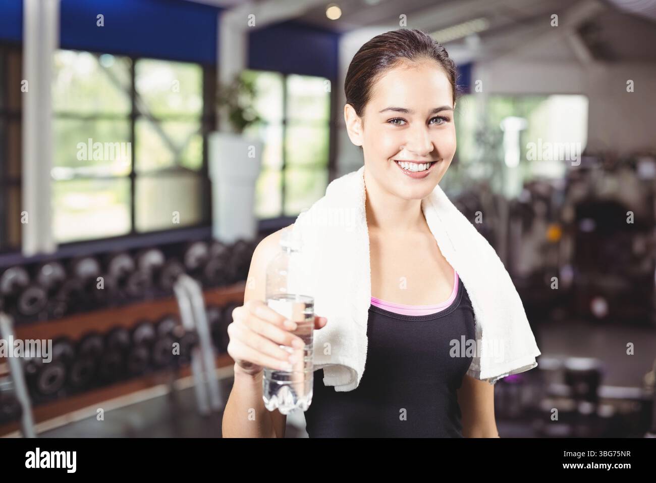 Donna che tiene in mano una bottiglia d'acqua e si trova in palestra con un'area pesi, con un pennarello e una pianta in vaso Foto Stock