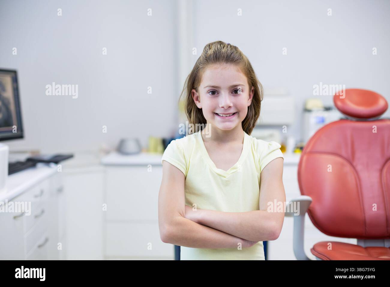 Bambino donna in piedi con le braccia incrociate in clinica odontoiatrica con sedia dentale rossa e monitor del computer Foto Stock