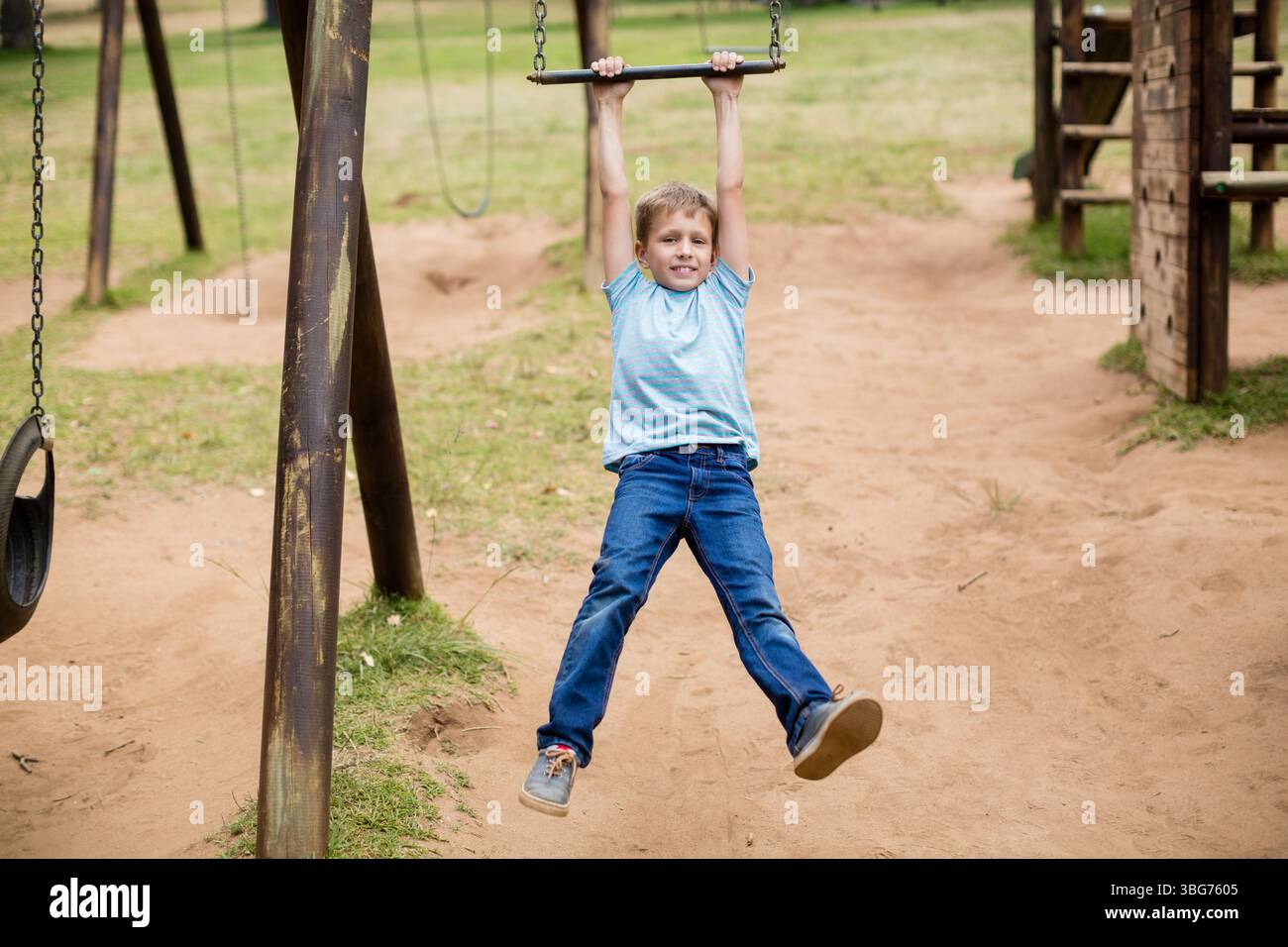 Ragazzo in età scolare appeso al bar orizzontale sul parco giochi con altalena e telaio in legno per arrampicate Foto Stock