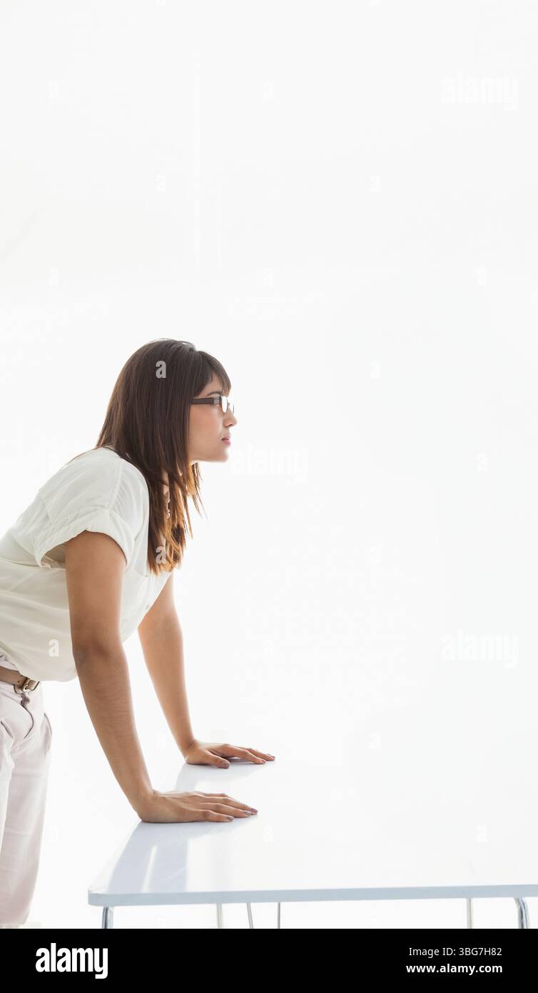 Donna asiatica che indossa occhiali appoggiati in avanti su una scrivania bianca in una luminosa camera minimalista che guarda a destra Foto Stock