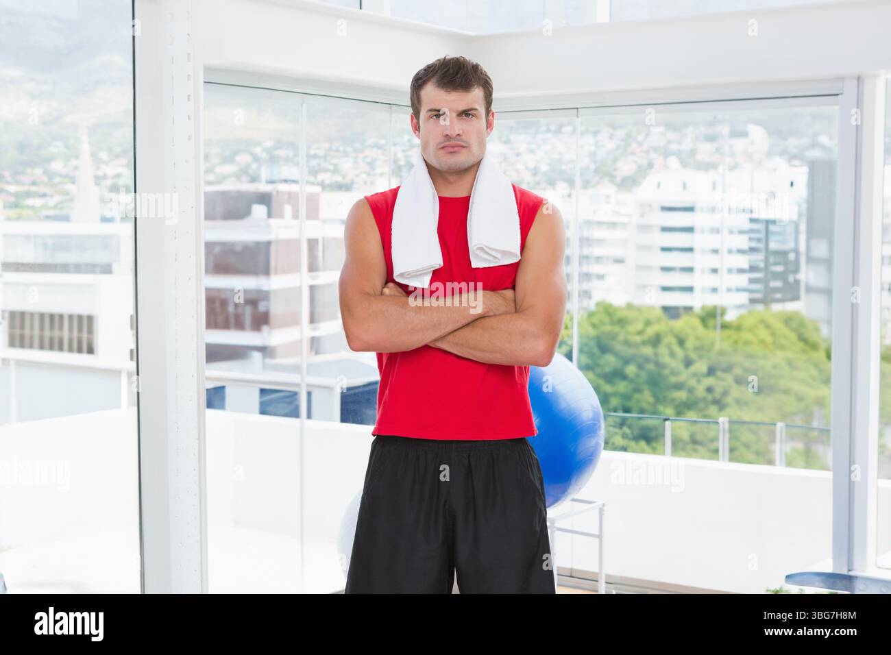 Uomo afroamericano in piedi accanto alle grandi finestre nella sala fitness con asciugamano e palla per esercizi Foto Stock
