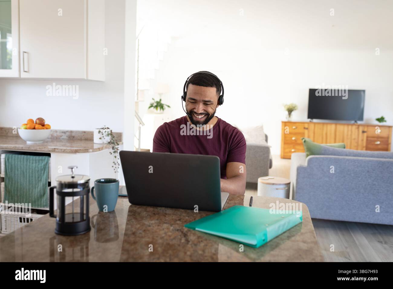 Uomo afroamericano che indossa le cuffie seduto a un'isola di cucina usando un computer portatile accanto alla stampa francese Foto Stock