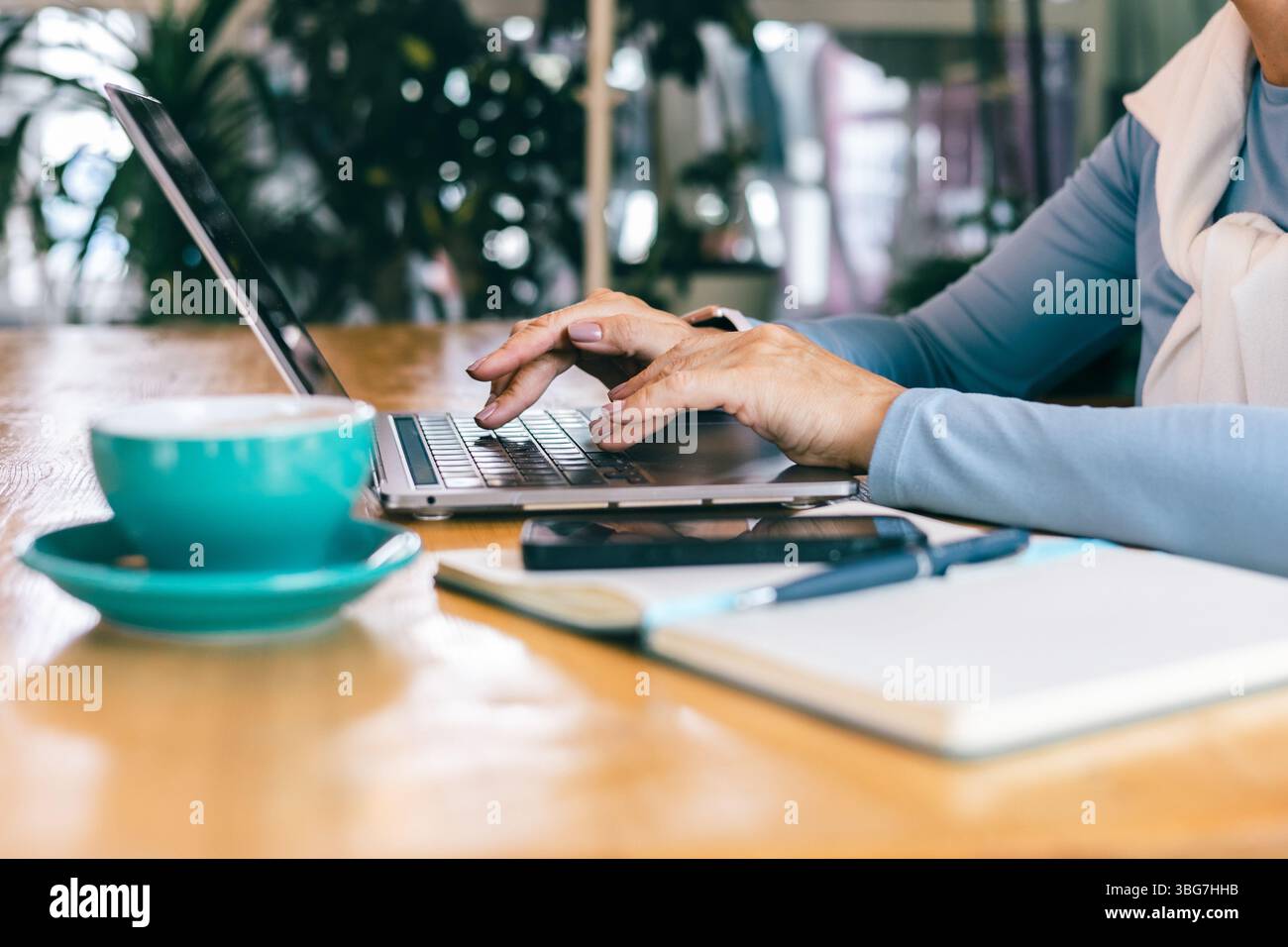 Le mani di una donna anziana che scrive su un computer portatile in un accogliente bar con notebook e caffè Foto Stock