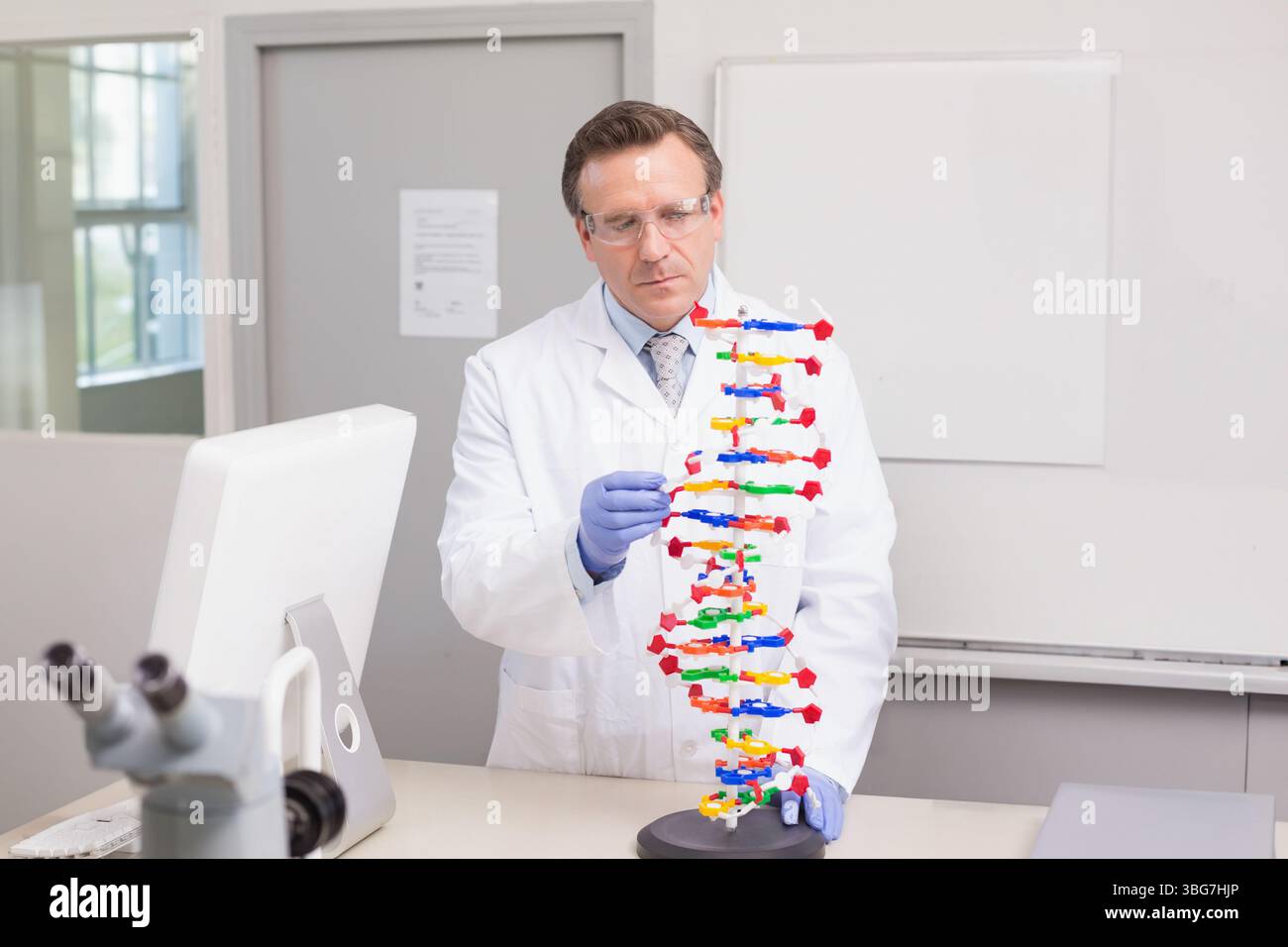Scienziato maschio che indossa guanti e occhiali di sicurezza che esamina il modello di DNA in plastica in laboratorio con microscopio Foto Stock