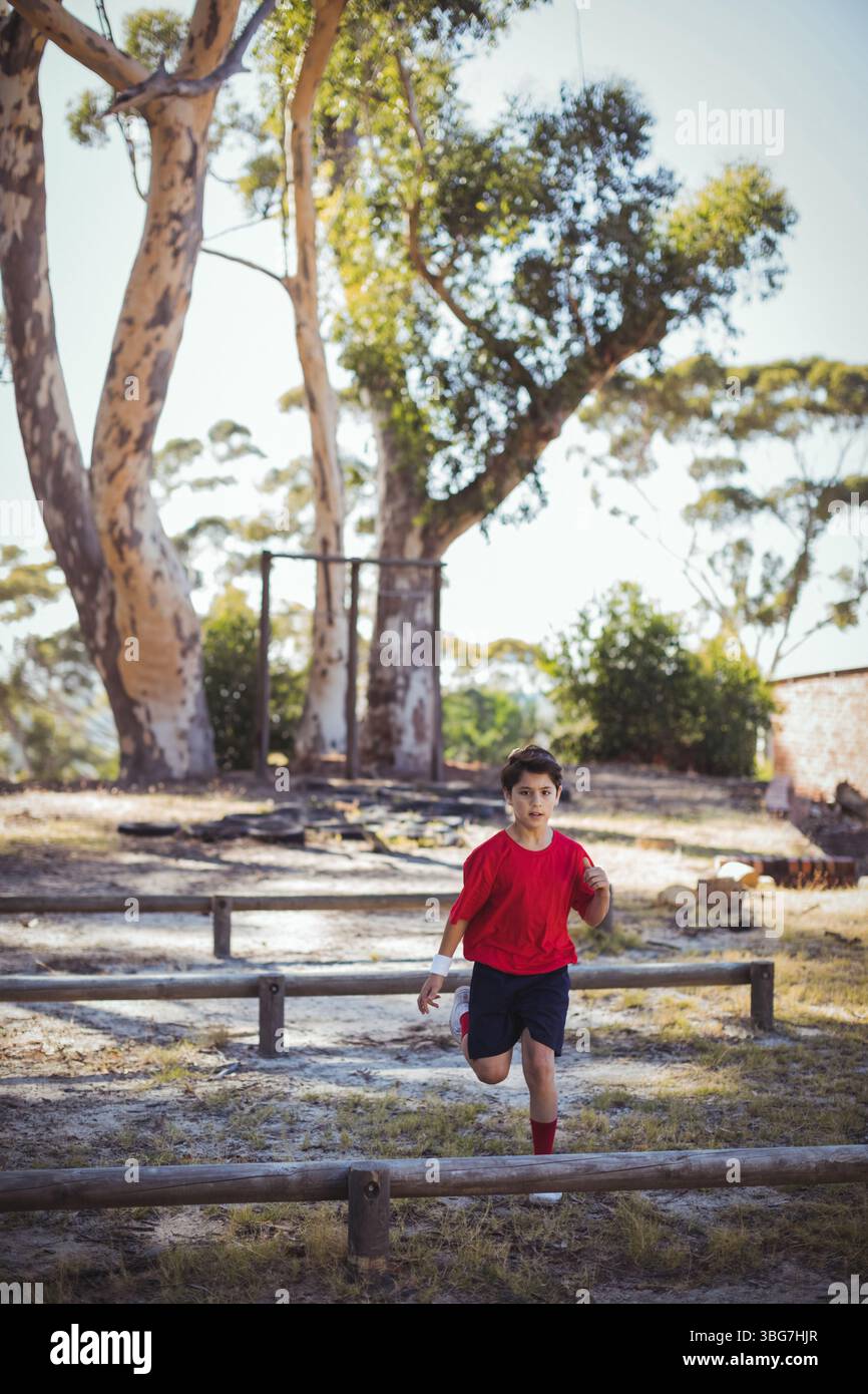 Ragazzo che corre lungo lo sgombero della sporcizia navigando in un percorso ad ostacoli con travi di legno che tiene la palla ovale nel parco Foto Stock
