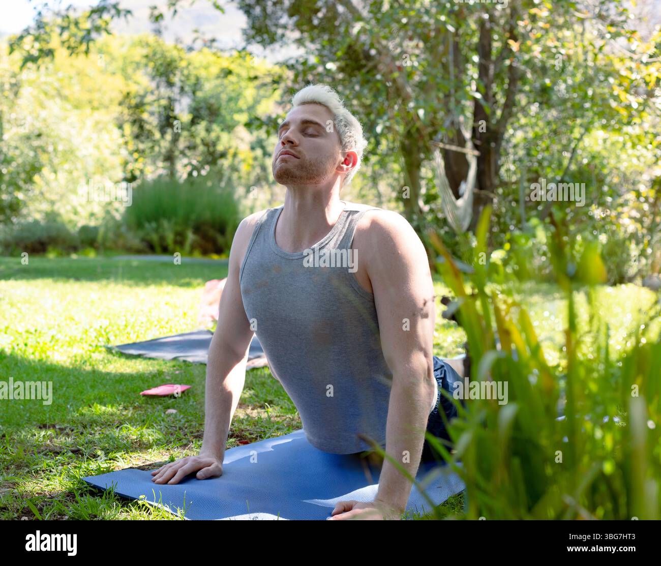 Uomo sulla trentina che pratica yoga in posa verso l'alto su un tappeto blu in un giardino illuminato dal sole con amaca Foto Stock