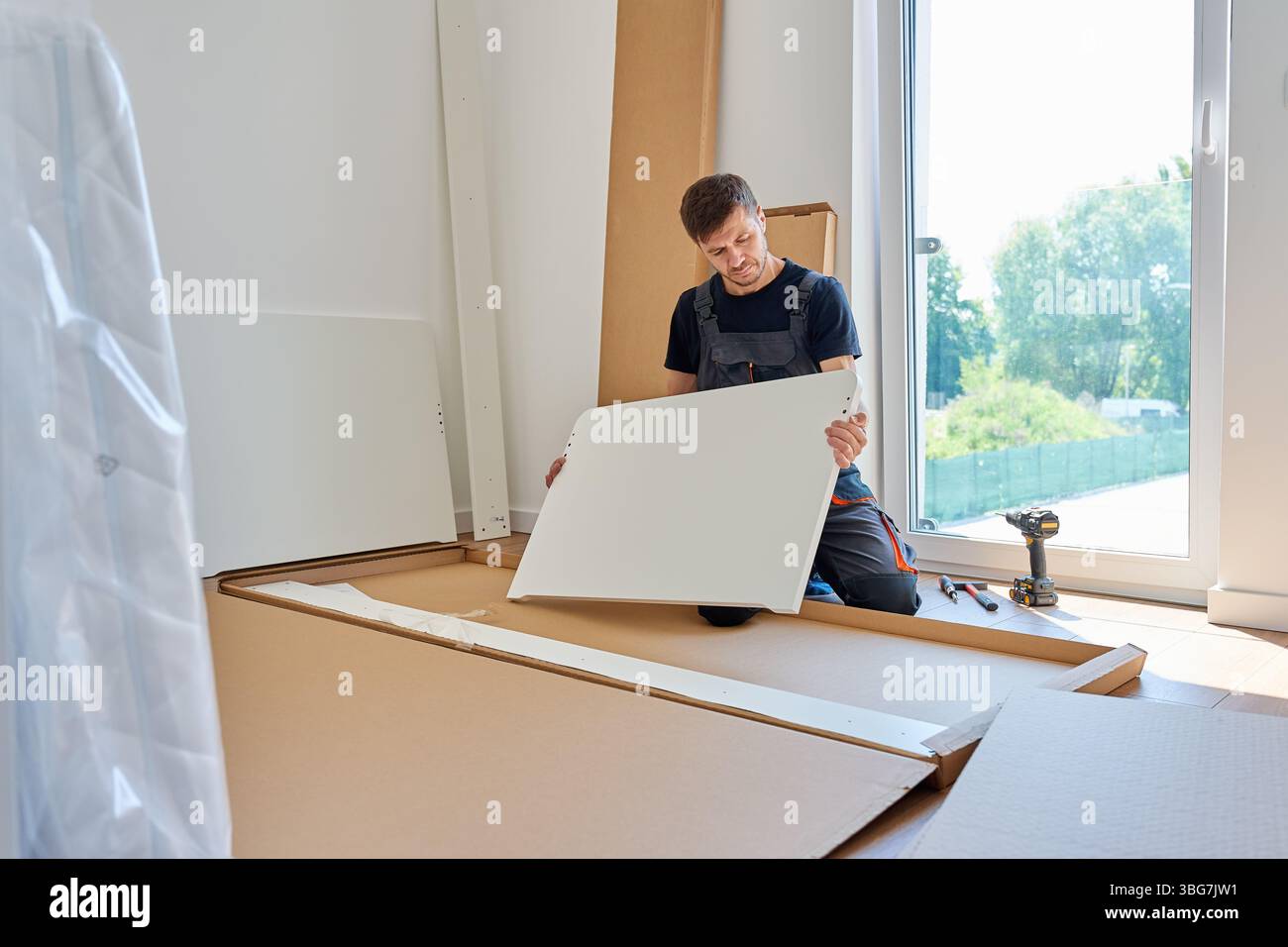 L'uomo in tuta da lavoro assembla un pannello di mobili bianco vicino a parti e utensili non imballati a casa. Tuttofare che prepara parte del letto per il montaggio in camera da letto Foto Stock