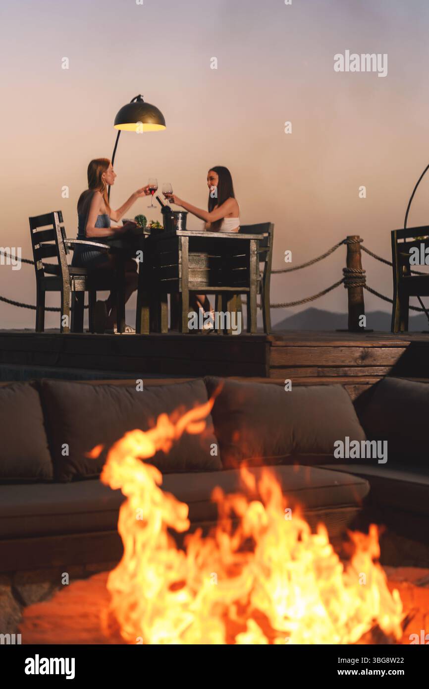 Una bruna e una bionda nel loro brindisi anni '20 con bicchieri da vino durante la cena, mentre le sfumature ardenti del tramonto dipingono il cielo dietro di loro. Foto Stock