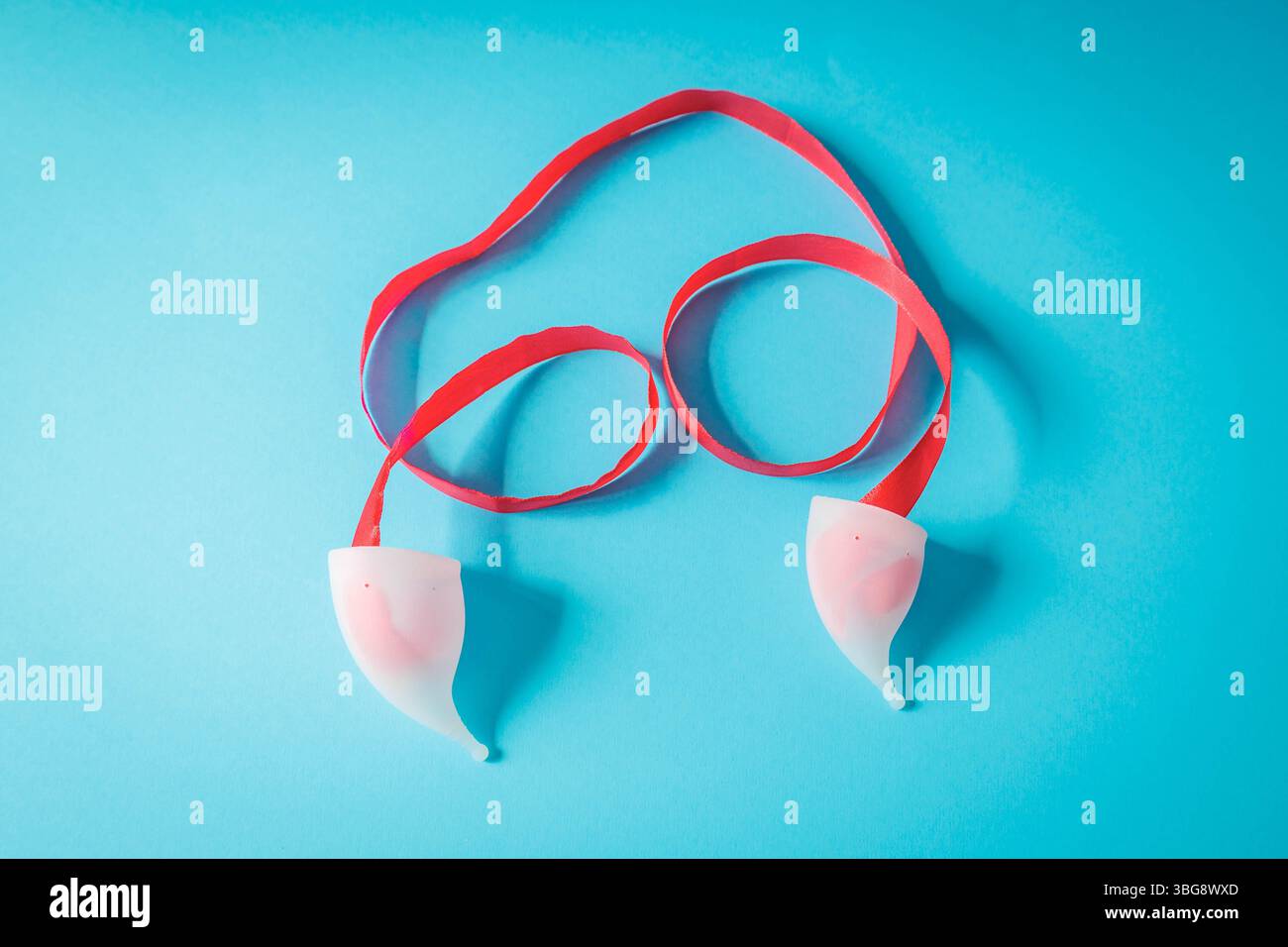 Coppette mestruali bianche con nastro rosso su sfondo blu, a forma di simboli di consapevolezza. Grafica creativa per la cura del periodo e le mestruali ecocompatibili Foto Stock