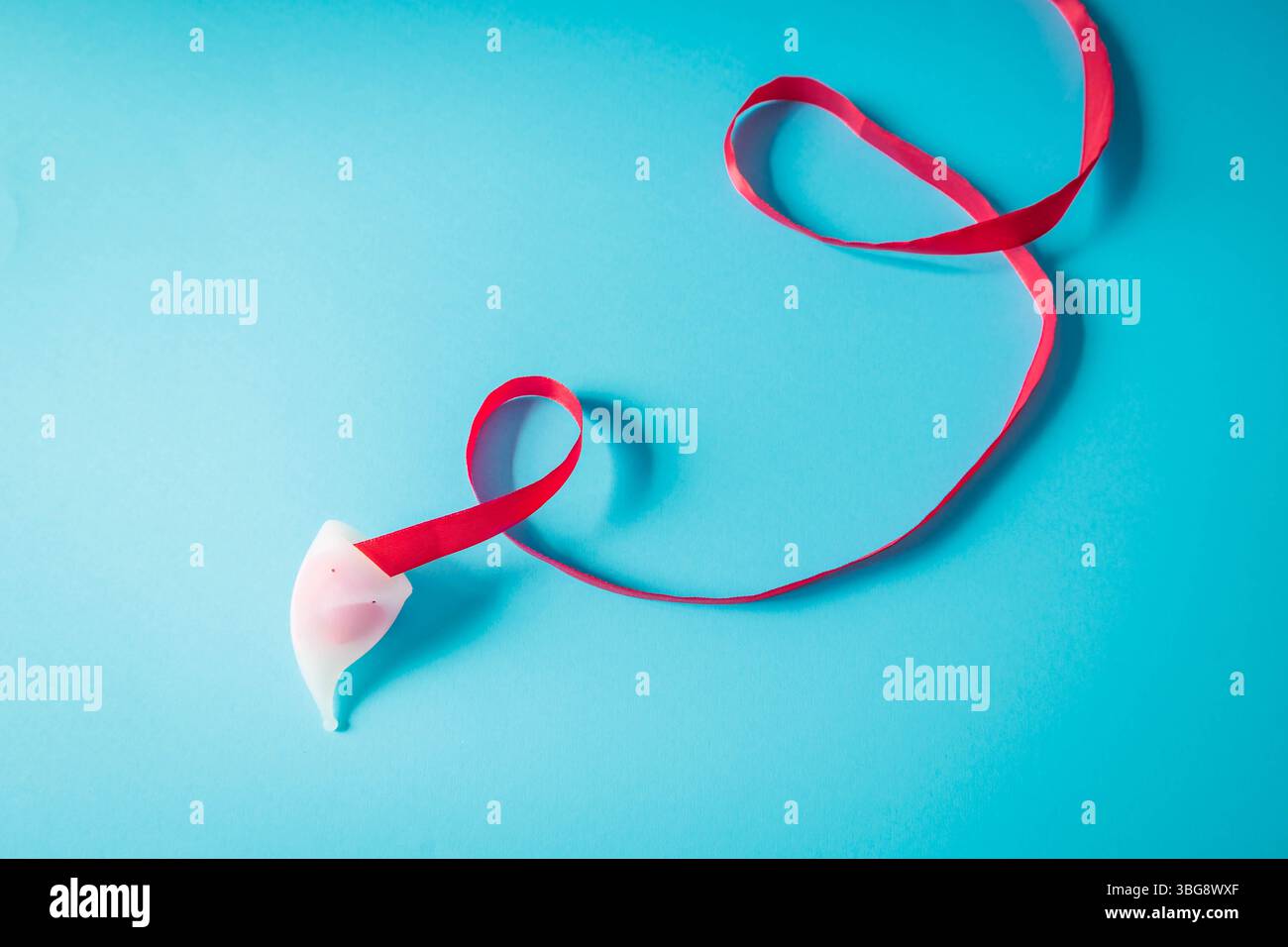 Composizione minimalista di una coppetta mestruale bianca con nastro rosso che scorre su sfondo blu. Immagine simbolica che promuove l'igiene femminile e l'ecocoscienza Foto Stock