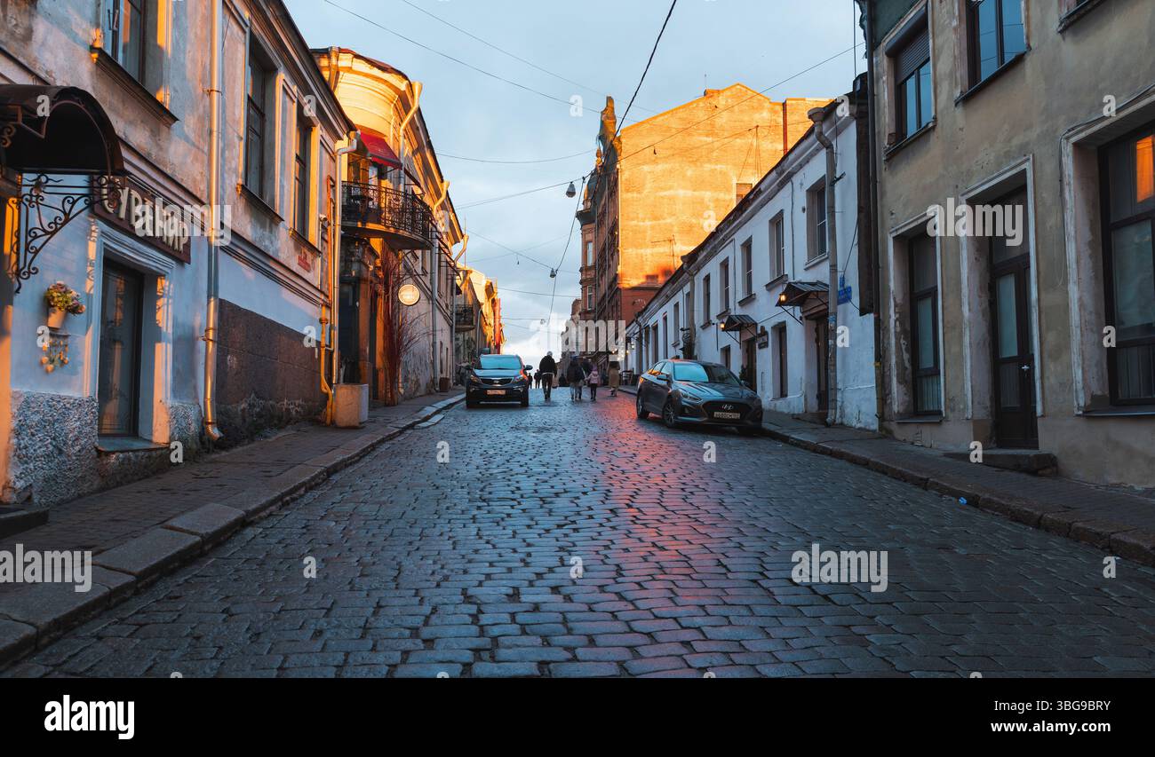 Vyborg, Russia - 3 maggio 2025: Affascinante strada acciottolata nella città vecchia di Vyborg illuminata dalla luce del sole dell'ora d'oro, che mostra auto parcheggiate, buil vecchio Foto Stock