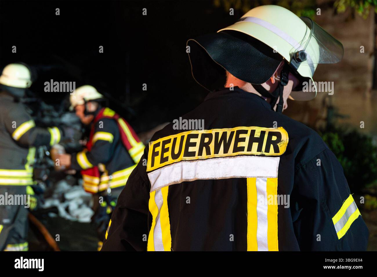LK Osnabrück, Deutschland 04. Juni 2025: IM Bild: Ein Feuerwehrmann, dessen Rücken mit dem markanten gelben Schriftzug FEUERWEHR versehen ist und der einen hellen Einsatzhelm trägt, blickt in die Szenerie eines nächtlichen Einsatzes. Seine Schutzkleidung ist mit gelben Reflexionsstreifen versehen. Im leicht unscharfen Hintergrund sind weitere Feuerwehrleute bei ihren Tätigkeiten zu erkennen, was die Teamarbeit und die koordinierte Vorgehensweise bei einem Feuerwehreinsatz unterstreicht Niedersachsen *** LK Osnabrück, Germania 04 giugno 2025 nella foto Un pompiere con il distintivo giallo Foto Stock