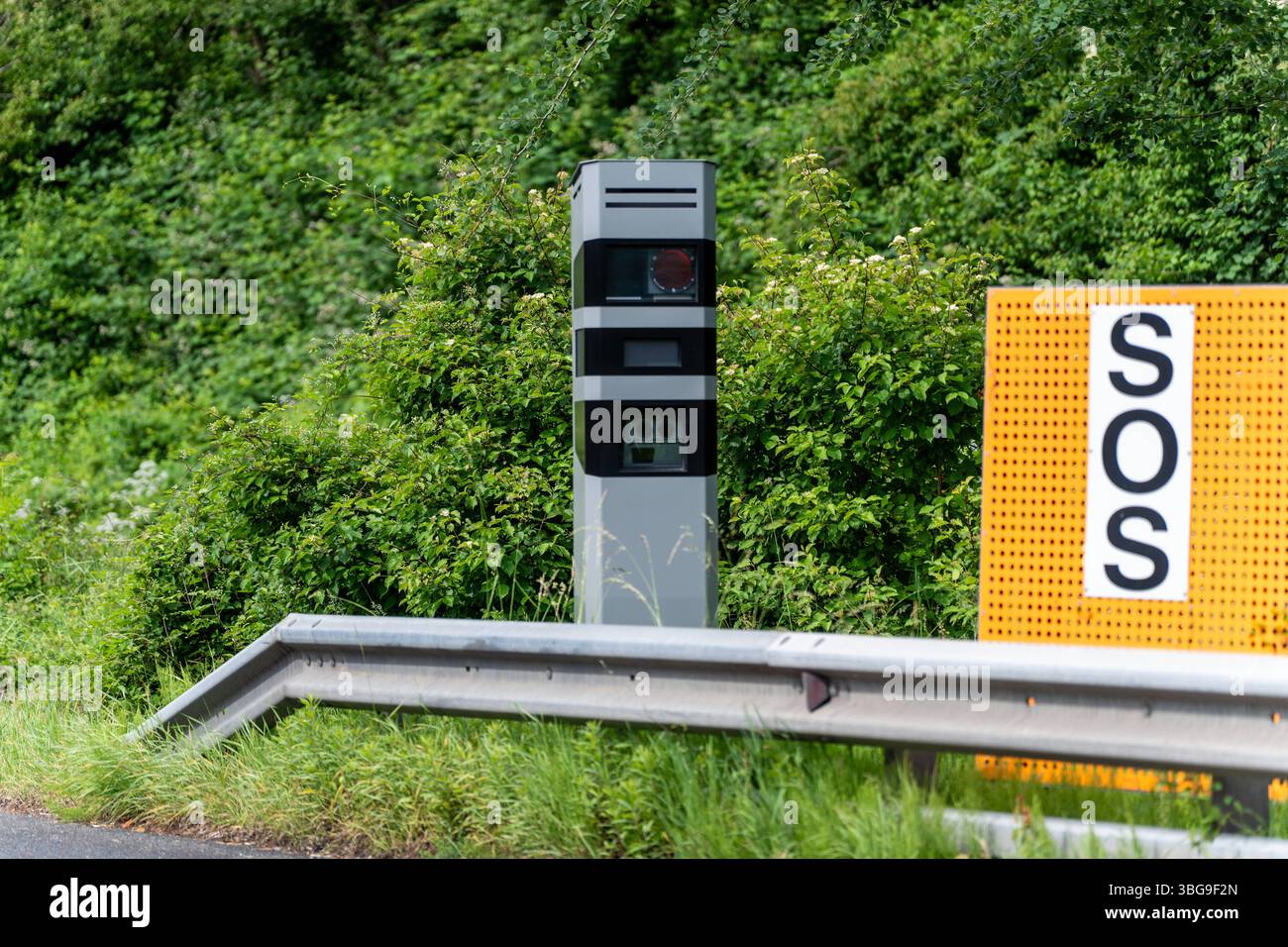 Vorarlberg, Austria - 4 giugno 2025: Un autovelox fisso accanto a un cartello SOS - una forte immagine simbolica dei rischi di velocità e delle conseguenze degli incidenti stradali o degli incidenti automobilistici causati dall'eccessiva velocità. *** Ein Fest installierter Blitzer neben einem SOS-Schild ein Starkes Symbolbild für die Risiken von Raserei und die Konsequenzen bei Verkehrsunfällen bzw. Autounfall durch überhöhte Geschwindigkeit. Foto Stock
