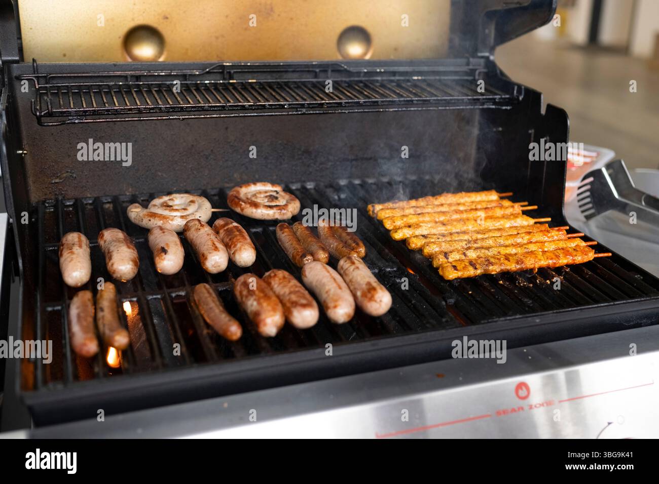 , Deutschland, 04.06.2025, Ein Gasgrill mit verschiedenen gegrillten Würsten, Spiralwürsten und gelb marinierten Fleischspießen. Rauch steigt auf – t Foto Stock