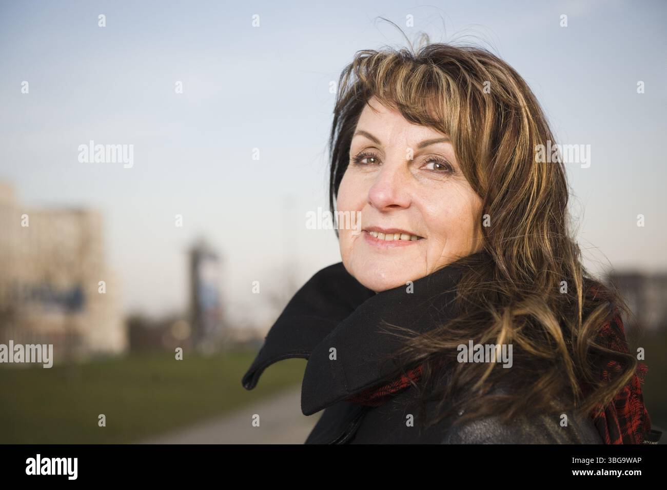 Ritratto testa e spalle di una donna di mezza età di fronte a uno sfondo urbano sfocato, sorridendo in modo amichevole nella fotocamera Foto Stock