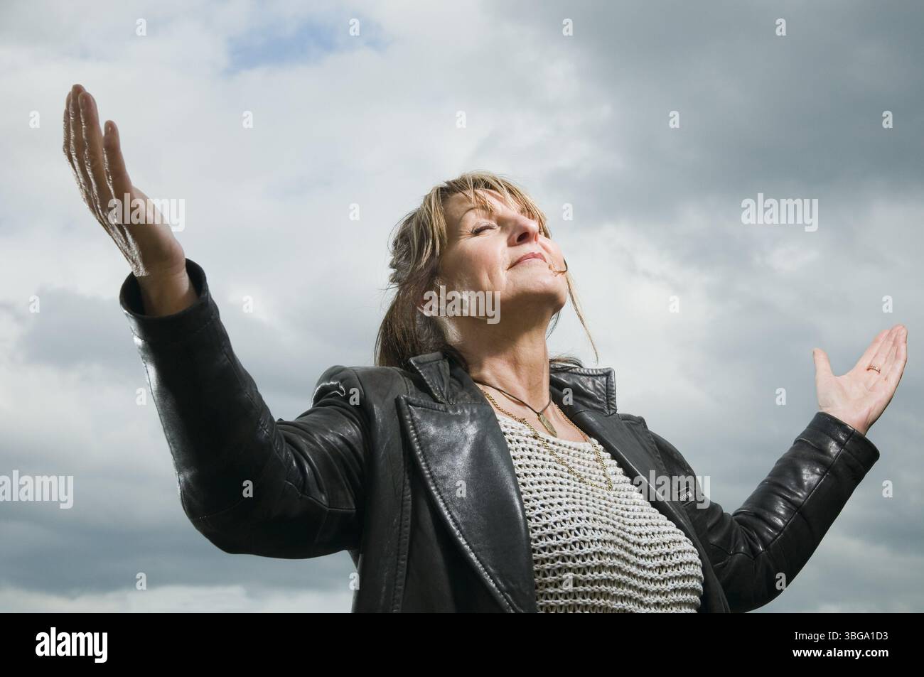 Vista della parte superiore del corpo di una donna di mezza età davanti a un cielo nuvoloso, che allarga le braccia e chiude gli occhi Foto Stock