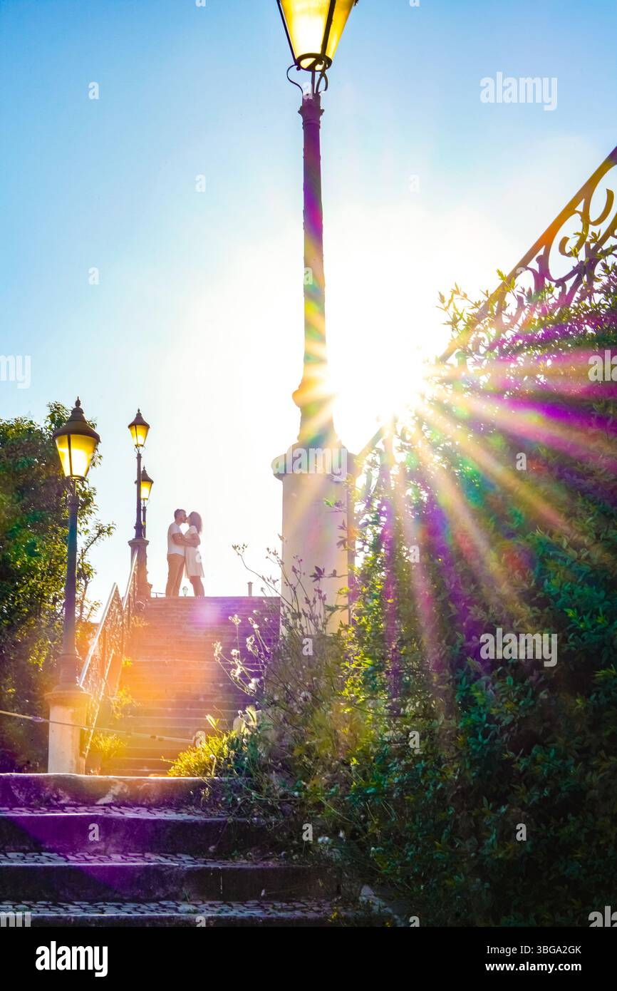Lisbona segreta: Una coppia romantica si trova su scale illuminate dal sole a Lapa, Lisbona, circondata da lampade vintage e vegetazione rigogliosa Foto Stock