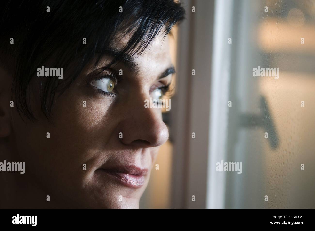 Ritratto laterale e oscuro della testa di una donna dai capelli neri di mezza età che sorride con attenzione e guarda fuori dalla finestra Foto Stock