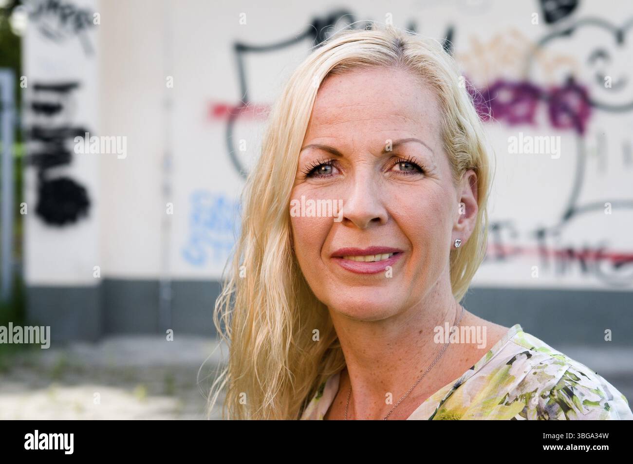 Ritratto frontale della testa e delle spalle di una donna bionda di mezza età dai capelli lunghi sorridente nella fotocamera di fronte a un muro di graffiti Foto Stock