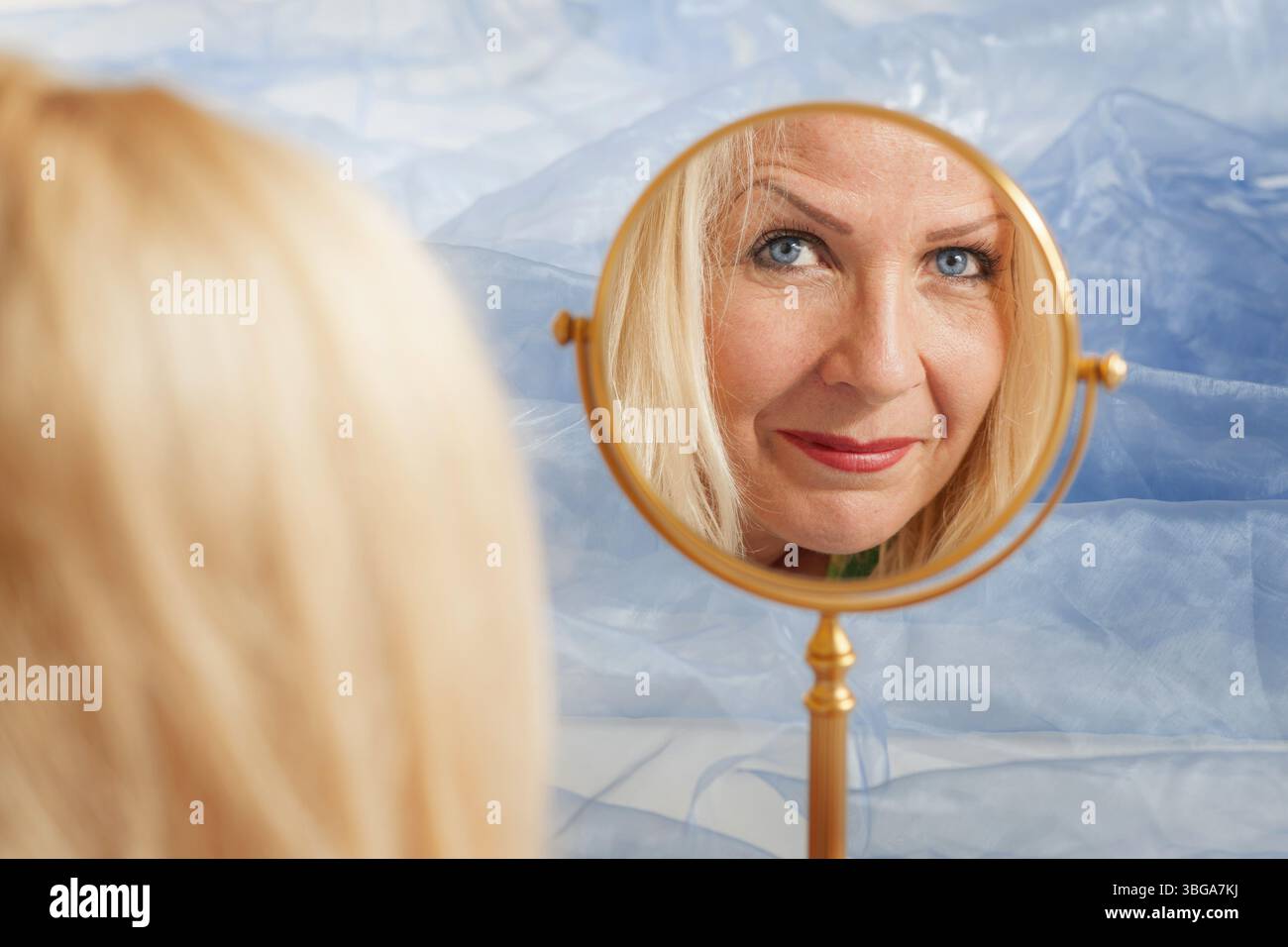 Riflesso del volto di una donna bionda di mezza età in uno specchio rotondo per il trucco su uno sfondo in tessuto rugoso azzurro Foto Stock