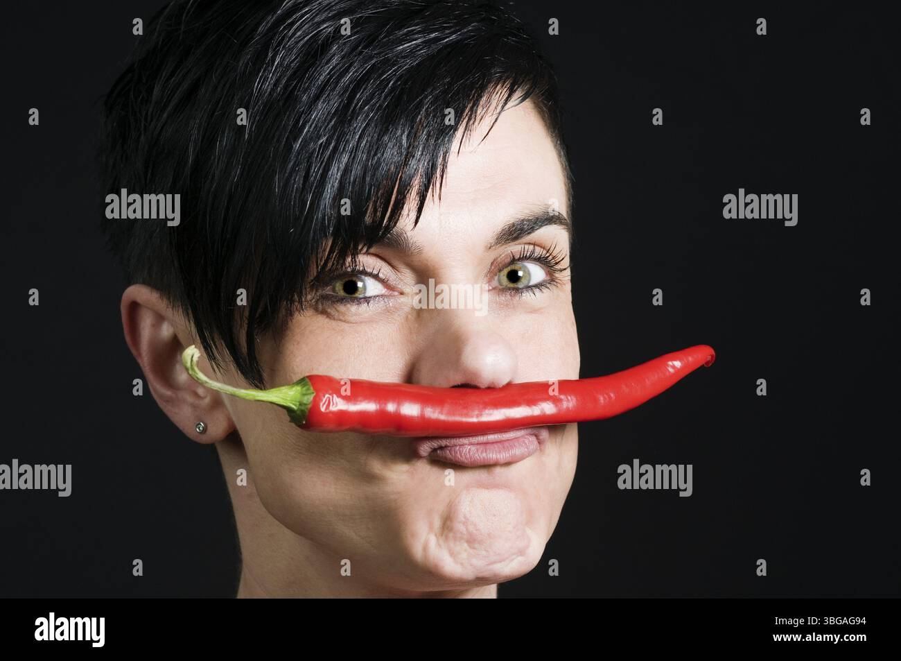 Ritratto della testa laterale di tre quarti di una donna dai capelli neri di mezza età su uno sfondo nero con un peperoncino stretto tra il labbro superiore e. Foto Stock