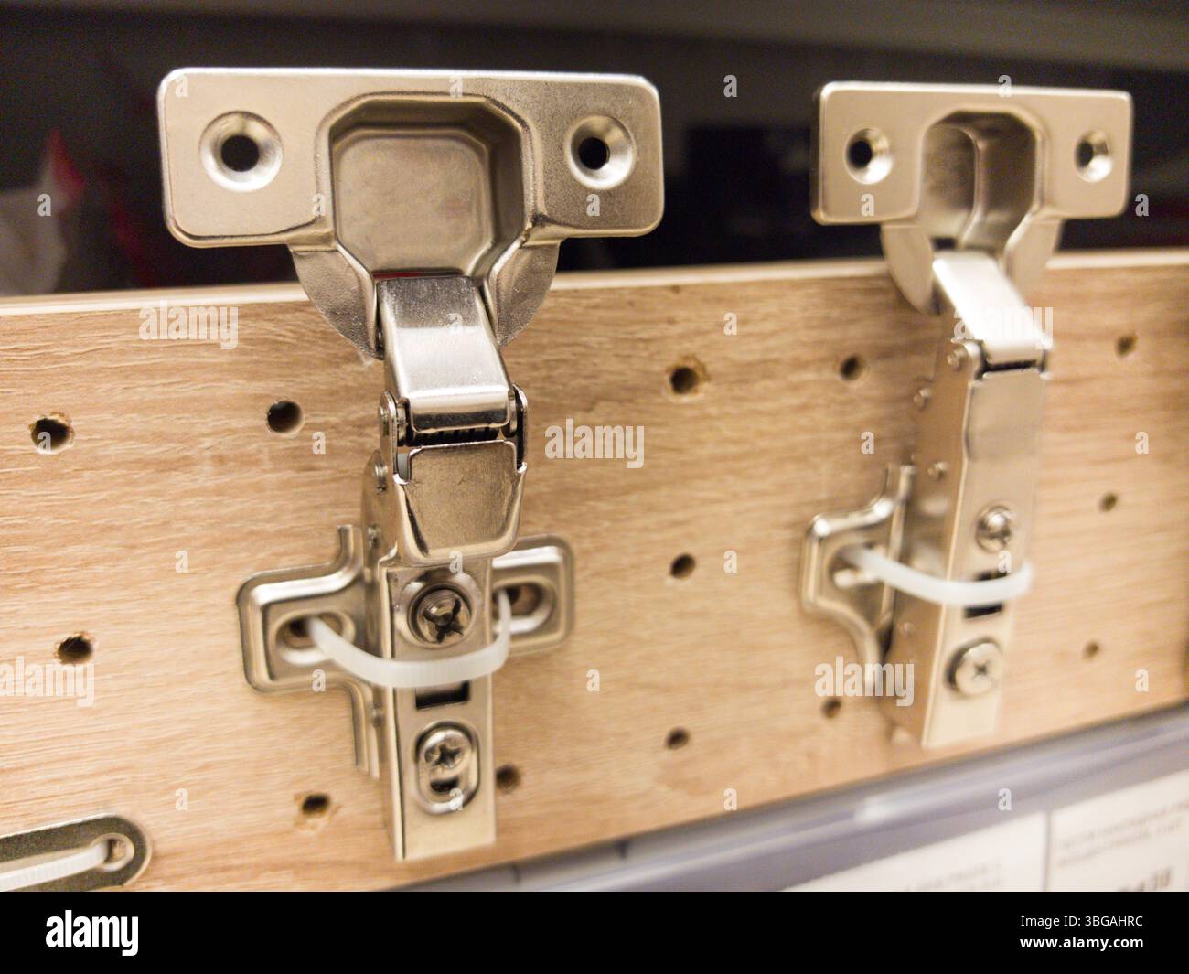 Le cerniere in metallo lucido per armadi sono montate su un pannello di legno che ne mostra il design. L'ambiente del negozio di hardware è luminoso e organizzato, perfetto per il fai da te Foto Stock