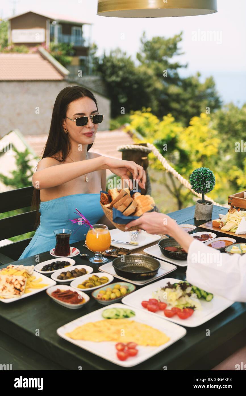 Una bruna anni '20 gusta la sua colazione al tavolo dell'hotel con splendide viste sul Mar Mediterraneo sullo sfondo. Foto Stock