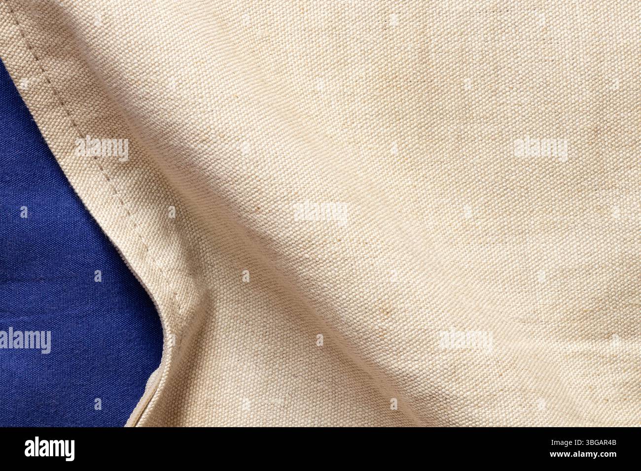 Tessuto in lino beige naturale come sfondo per design e creatività con una piega e parte del tessuto blu sottostante, sfondo in tessuto Foto Stock