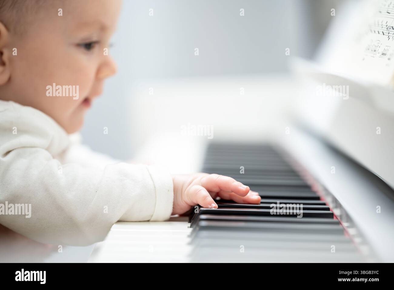 Descrizione: Vista laterale di un bambino in un maglione bianco che suona il pianoforte in una stanza luminosa. Vita giovane e musica. Foto Stock