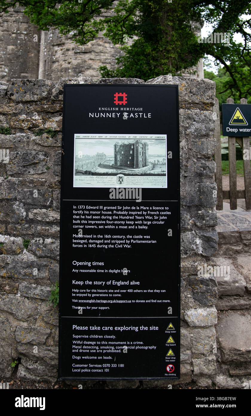 Cartello informativo sul castello di Nunney, un sito patrimonio dell'umanità inglese, esposto su un muro di pietra con vegetazione sullo sfondo Foto Stock