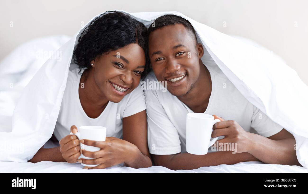 Felice coppia nera che posa sotto coperta, bere tè Foto Stock