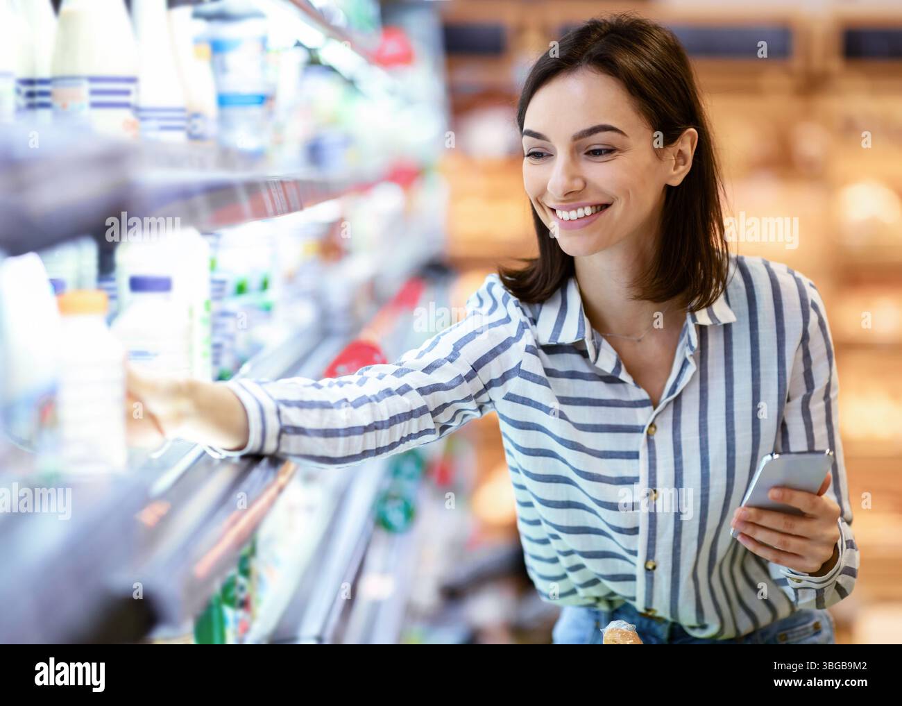 Donna che tiene lo smartphone in piedi in negozio prendendo il latte Foto Stock