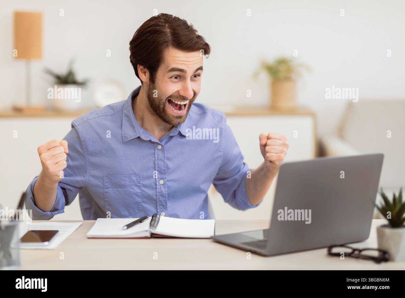 Eccitato giovane uomo d'affari al computer portatile scuotendo i Fists in ufficio Foto Stock