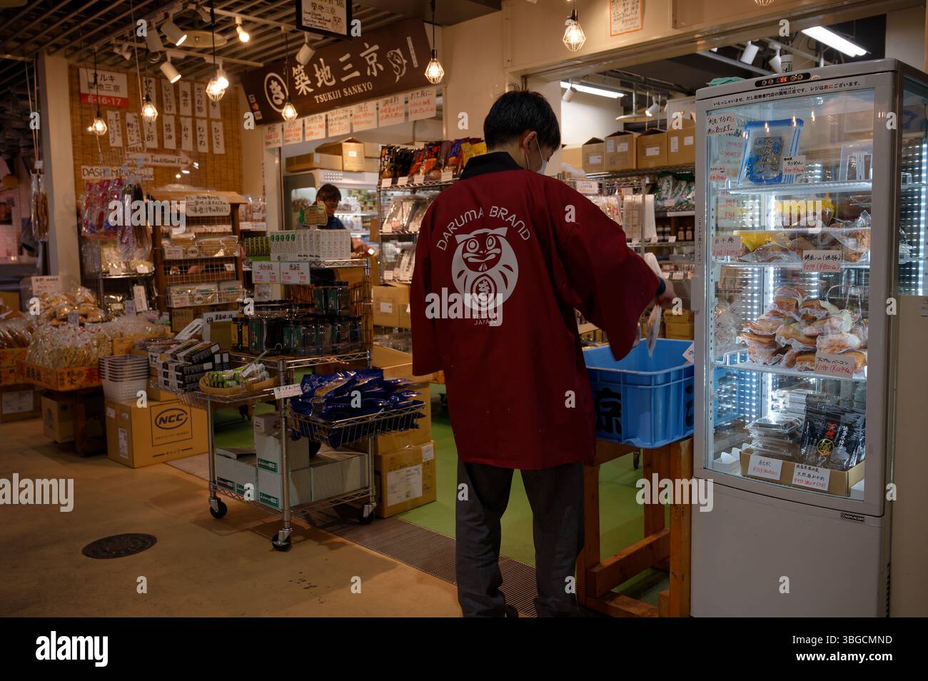 Il negoziante in un cappotto Daruma Brand rifornisce pesce secco in un vivace negozio di Tsukiji Market Foto Stock