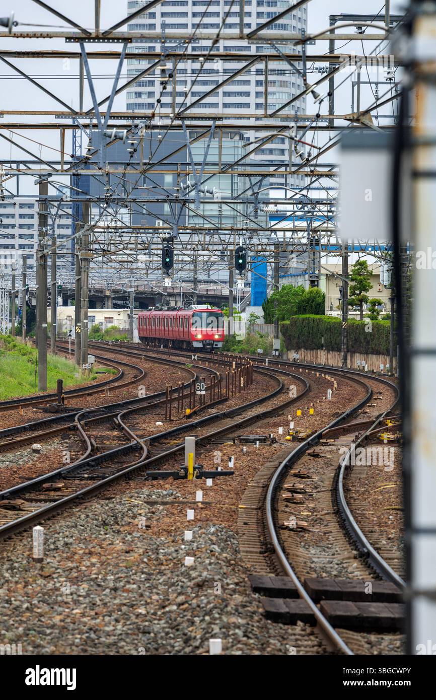 Il treno rosso per pendolari viaggia lungo binari curvi in ambiente urbano. Circondati da edifici alti, i binari sono fiancheggiati da linee elettriche aeree. Foto Stock