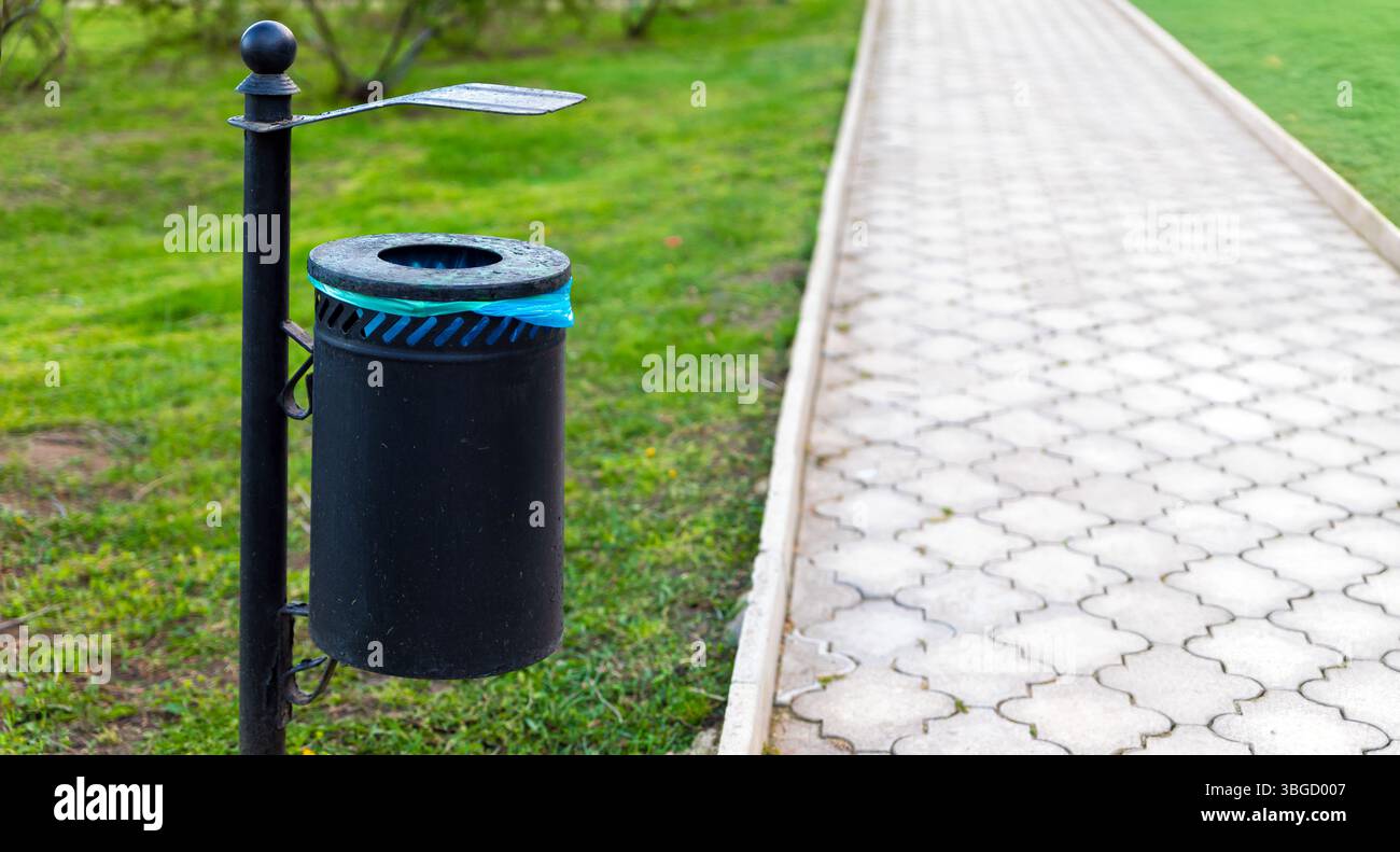 Cestino in metallo nero con rivestimento blu montato accanto a un passaggio pavimentato a motivi geometrici in un parco pubblico verde. Foto Stock