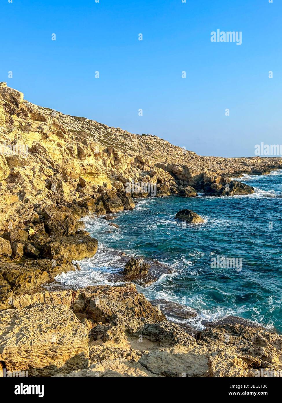 La luce del sole dorata bagna un'aspra costa rocciosa mentre le onde si infrangono contro la costa sotto un cielo azzurro. Un paesaggio marino sereno e selvaggio perfetto per la natura - Immagine stock catturata con smartphone