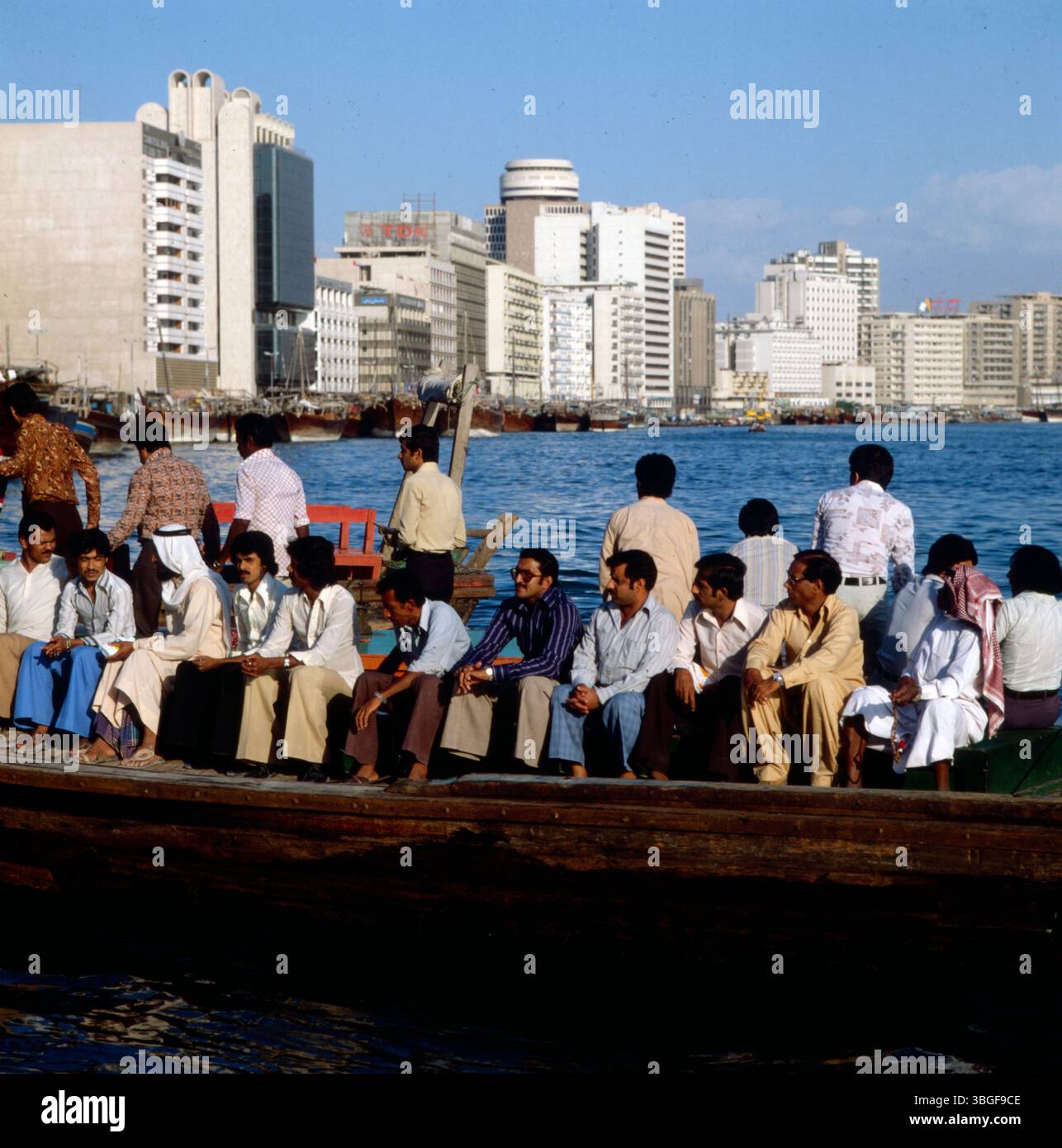 Pendler auf dem Weg zur Arbeit auf einem Holzboot vor der Uferbebauung von Dubai. Im Hintergrund moderne Hochhäuser entlang der Wasserkante. Foto Stock